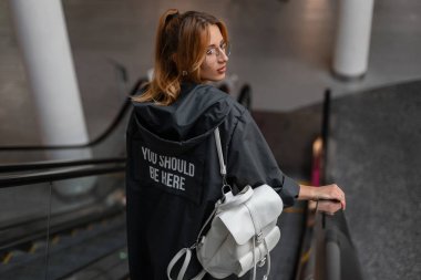 Gözlüklü beyaz deri sırt çantası ile şık bir ceket çekici genç kadın hipster mağazada yürüyen merdiven üzerinde duruyor. Trendy güzel kız dükkan da seyahat.