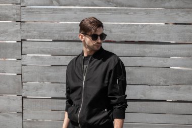 Bir saç modeli ile siyah şık güneş gözlüğü şık siyah giysiler içinde Çekici genç adam hipster şehirde gri ahşap duvar yakınında duruyor. Yakışıklı kentsel adam esmer açık havada. Yaz güneşli gün