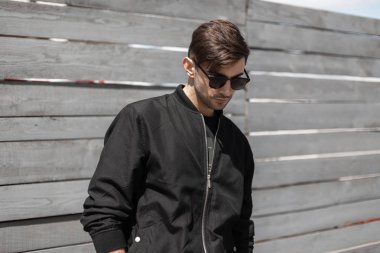 Modaya uygun koyu güneş gözlüğü şık siyah ceket moda saç modeli ile Genç yakışıklı hipster adam güneşli bir yaz gününde ahşap bir vintage duvar yakınında pozlar. Amerikalı esmer adam açık havada.