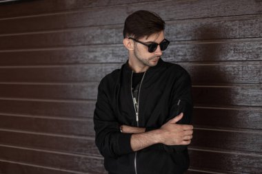 Modaya uygun bir saç stilinde şık güneş gözlüğü şık siyah ceket cool genç hipster adam sokakta bir vintage kahverengi ahşap binanın yakınında dinleniyor. Modern seksi adam kapalı poz. Sokak stili.