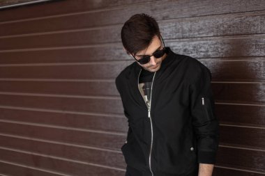 Modaya uygun koyu güneş gözlüğü şık siyah giysiler şık saç modeli ile Kentsel genç hipster adam sokakta kahverengi bir vintage ahşap duvar yakınında dinleniyor. Seksi erkek modeli. Sokak modası.