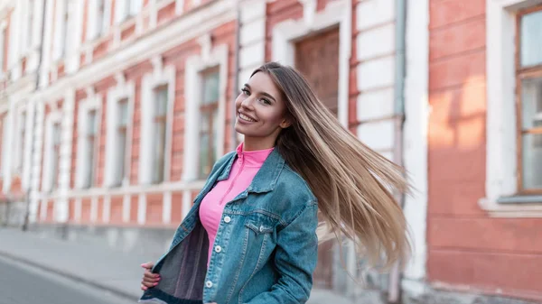 Lebenslustige junge Frau mit süßem Lächeln und blonden langen Haaren in einer Vintage-Jeansjacke in einem trendigen rosafarbenen Top posiert in der Nähe eines Gebäudes auf der Straße. ziemlich glückliches Mädchen spaziert an einem Sommertag durch die Stadt. — Stockfoto