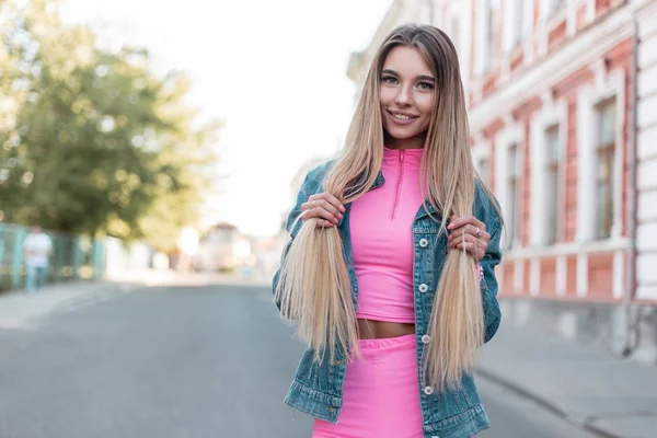 Позитивная молодая женщина с красивой милой улыбкой в модном джинсовом пиджаке в стильной розовой рубашке в шортах стоит на дороге в городе и держит волосы. Привлекательная блондинка позирует . — стоковое фото