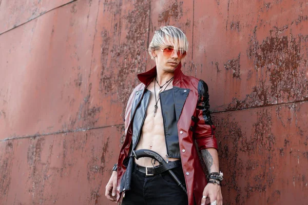 Stijlvolle jonge hipster man met blond haar in glazen in een leren jas in jeans met een vintage pistool poses in de buurt van een roestige muur. Sexy vent buitenshuis. — Stockfoto