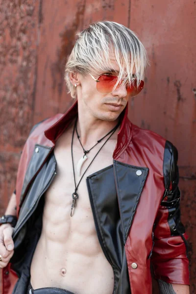 Крутой молодой блондин с голым туловищем в модном красно-черном кожаном пиджаке в винтажных красных солнцезащитных очках позирует возле старого ржавого здания . — стоковое фото