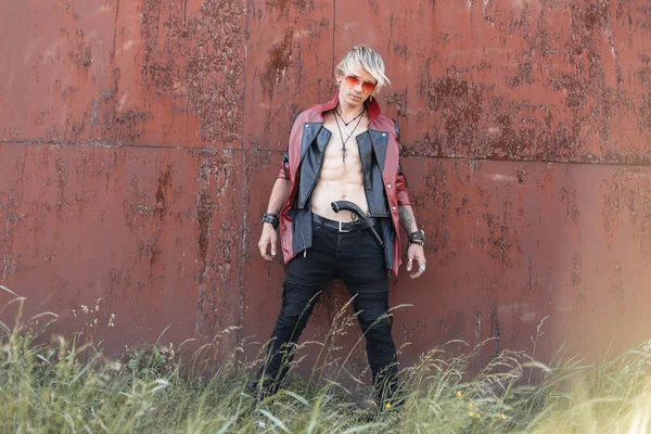 Старомодный молодой хипстер со светлыми волосами в очках в красно-черной кожаной куртке в джинсах со старым оружием позирует возле ржавой стены в яркий летний день. — стоковое фото