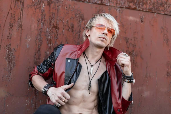 Сексуальный спортивный модный молодой человек хипстер в стильной черно-красной кожаной куртке в красных очках с модными позами для волос возле старинной ржавой стены — стоковое фото