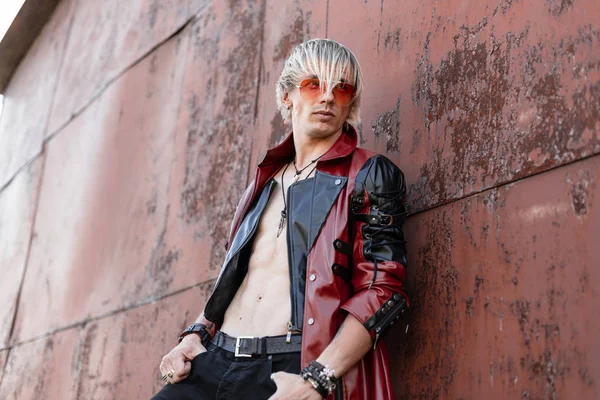 Sexy Europese jonge hipster man in rode zonnebril in een zwart vintage lederen jas met een naakte torso in jeans ontspant in de buurt van een oude roestige metalen wand op straat. — Stockfoto