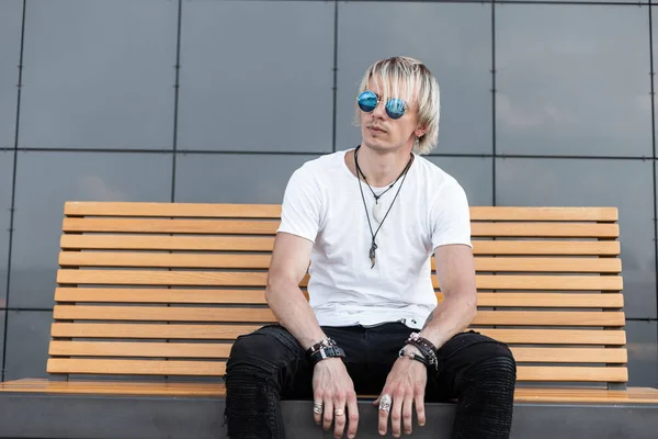 Young nice hipster man in blue sunglasses in black jeans in white t-shirt sit on a vintage wooden bench near a modern gray wall in the street. Handsome guy model outdoors. Stylish summer menswear. — Stock Photo, Image
