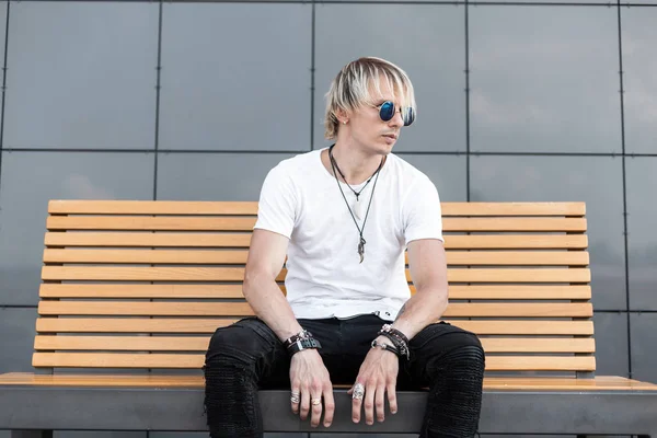 Auf einer Holzbank in der Stadt sitzt ein junger attraktiver Hipster mit modischer Frisur, trendiger Sonnenbrille und stylischem T-Shirt in Jeans. gutaussehender blonder Kerl entspannt im Freien. Sommer-Stil. — Stockfoto