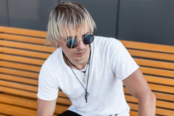 Der hübsche junge Hipster-Mann im schicken weißen T-Shirt mit stylischer blauer Sonnenbrille und Amuletten am Hals sitzt draußen auf einer Holzbank. Der amerikanische Stadtmensch ist blond und entspannt in der Stadt. Straßenstil — Stockfoto