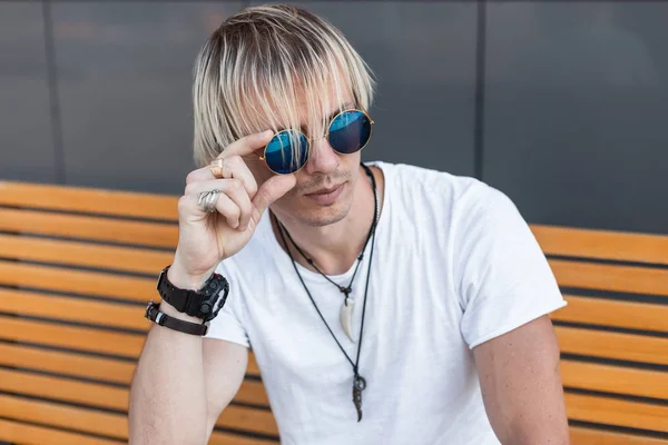 Young cool hipster man blond in a trendy white t-shirt with vintage amulets around neck sits on a yellow wooden bench and straightens stylish blue sunglasses. Handsome guy is relaxing in the city. — Stock Photo, Image