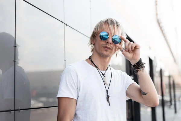 El joven hombre hipster de moda con elegantes camisetas blancas endereza las gafas de sol vintage al aire libre. Cool chico americano rubio se relaja en la ciudad en un día de verano brillante. Moda callejera. Hora de verano — Foto de Stock