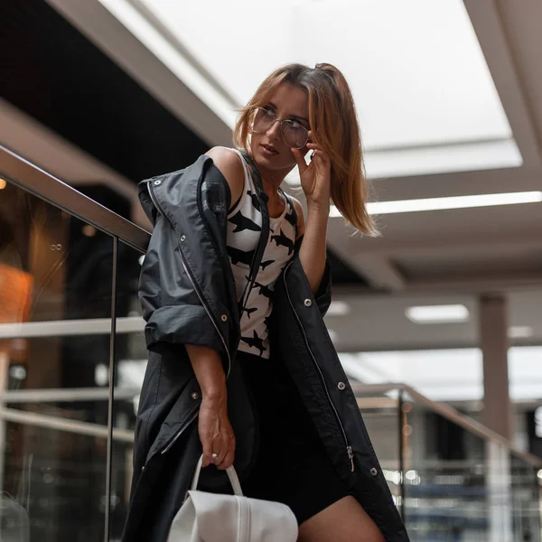 Mulher bonita em uma jaqueta vintage em uma camiseta com óculos em uma saia com uma mochila de couro está de pé perto da parede de vidro em uma loja moderna. Modelo de menina atraente de cabelos vermelhos na moda . — Fotografia de Stock