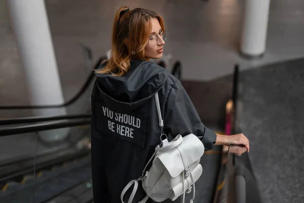 Atractiva joven hipster mujer en una chaqueta elegante con un patrón con una mochila de cuero blanco con gafas está de pie en la escalera mecánica en la tienda. Moda hermosa chica viaja a través de la tienda . — Foto de Stock