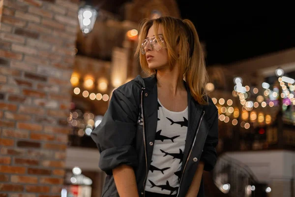 Jolie jolie jeune femme hipster aux cheveux roux dans des lunettes élégantes dans un t-shirt dans une veste à la mode est debout dans une histoire moderne et profiter du week-end. Européen belle fille sur une promenade . — Photo