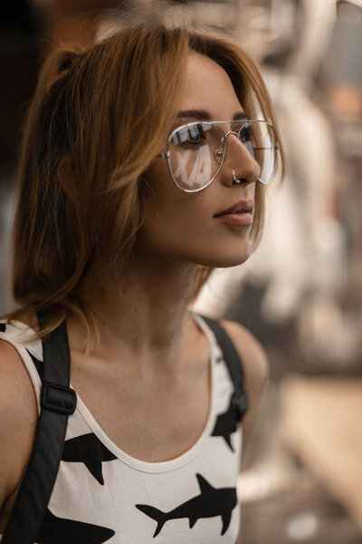 Portrait of a beautiful young hipster woman in trendy glasses with an earring in nose with a stylish hairstyle in a fashionable t-shirt with a pattern near the glass showcase in a store. Pretty girl.