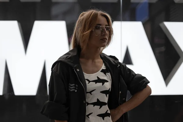 Mujer joven bastante hermosa de moda en ropa de moda en gafas elegantes está de pie cerca de un escaparate de cristal moderno en un centro comercial. Atractiva chica hipster pelirroja en el interior. Moda juvenil . — Foto de Stock