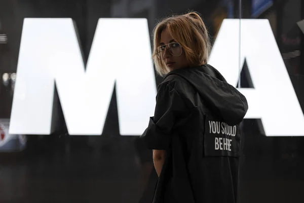 Joven mujer pelirroja hipster en gafas de moda en una chaqueta de moda está de pie en un centro comercial cerca de un escaparate de cristal brillante. Hermosa modelo urbana chica de vacaciones. Moda juvenil . — Foto de Stock