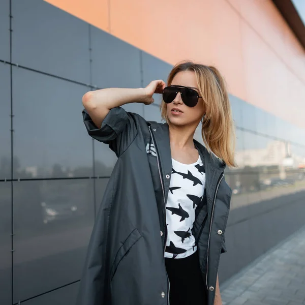 Jolie jolie jeune femme hipster en vêtements élégants dans des lunettes de soleil noires à la mode pose dans une ville près d'un bâtiment gris moderne. Européen fille aux cheveux roux bénéficie d'une journée ensoleillée brillante . — Photo