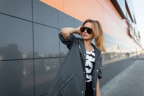 Jeune femme urbaine hipster aux cheveux roux dans une veste à la mode en lunettes de soleil noires dans un t-shirt blanc avec un motif posant à l'extérieur près d'un mur moderne. Jolie fille dans la ville dans une journée d'été lumineuse — Photo