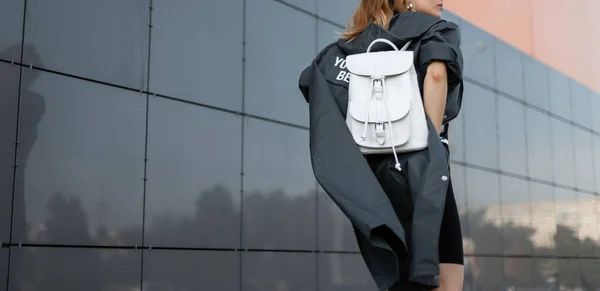 Visão traseira em uma jovem mulher de moda em uma jaqueta de moda longa com uma mochila de couro branco elegante andando pela rua perto do edifício moderno. Roupas femininas de estilo americano. Close-up . — Fotografia de Stock