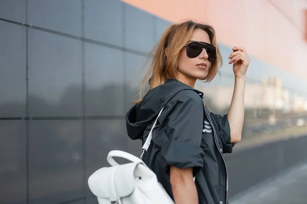 Roodharige mooie jonge hipster vrouw in stijlvolle jas met vintage lederen rugzak in trendy zonnebrillen staan in de buurt van modern gebouw in de stad op een zomerdag. Urban mooi meisje model. — Stockfoto