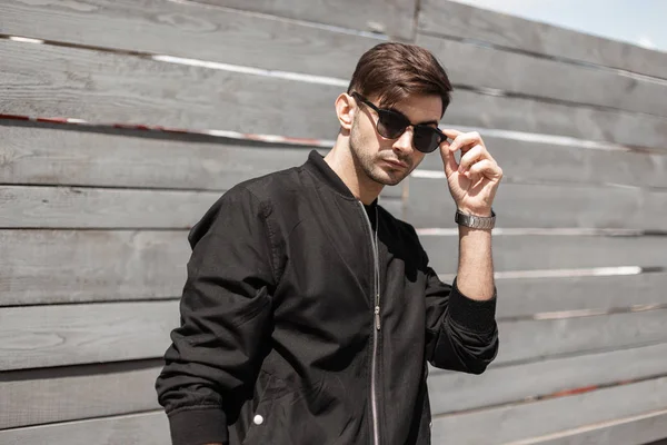 Urban American hipster young man in a black stylish jacket stands and straightens trendy sunglasses near a wooden fence on a bright summer sunny day. Handsome sexy guy brunette outdoors. — Stock Photo, Image