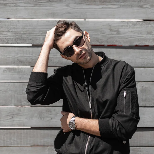 Modern handsome young man in trendy black jacket in stylish sunglasses with a hairstyle poses near a vintage wooden wall on the street. Nice hipster guy relaxes on a summer day on the street. Menswear — Stock Photo, Image