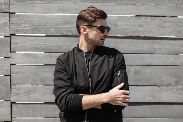Cool American young hipster man in stylish dark sunglasses in trendy black jacket poses near a vintage wooden wall on the street. Attractive modern guy with a fashionable hairstyle. Menswear. — Stock Photo, Image