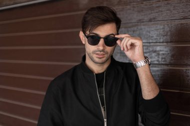 Bir saat ile şık bir saç modeli ile siyah bir ceket trendy güneş gözlüğü yakışıklı seksi genç hipster adam kapalı ahşap bir duvar yakınında pozlar. Yakışıklı adam. Moda erkek giyim ve aksesuarları.