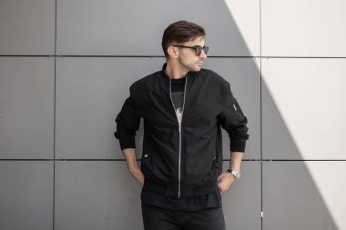 Şık bir ceket güneş gözlüğü yakışıklı modern genç adam şehirde sokakta modern bir gri duvar yakınında duran rahatlatır. Çekici adam hipster açık havada parlak yaz güneşi sahiptir.