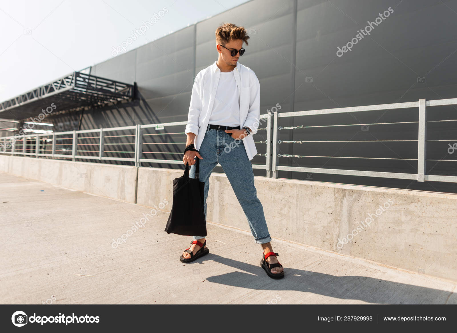 穿着时尚服装、戴着太阳镜、黑色布袋、穿着复古凉鞋的年轻人,在一座灰色建筑附近的城市里。美国人时髦的男士夏季服装。青年风格. ——图库图片©, image size:1600x1167