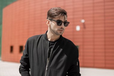 Şık saç modeli ile siyah trendy ceket şık güneş gözlüğü çekici genç hipster adam bir yaz gününde şehir etrafında yürür. Modern binanın yakınındaki yakışıklı adam. Amerikan tarzı.