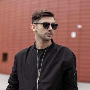 Kırmızı modern binanın yakınında sokakta ayakta Vintage güneş gözlüğü trendy ceket şık bir saç modeli ile yakışıklı moda genç hipster adam. Çekici adam şehirde yürür.