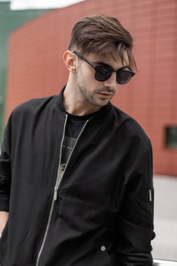 Trendy saç tarzı ile şık güneş gözlüğü moda ceket Amerikan güzel genç hipster adam bir yaz gününde şehirde yürür. Açık havada kırmızı bina yakın yakışıklı adam modeli. Sokak stili.