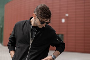 Kırmızı modern binanın yakınında sokakta duran karanlık güneş gözlüğü siyah trendy ceket şık bir saç modeli ile genç yakışıklı moda hipster adam. Çekici adam şehirde yürür.