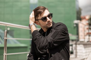 Şık siyah ceket Vintage güneş gözlüğü Trendy saç ile yakışıklı genç esmer adam güneşli bir yaz gününde şehirde dinleniyor. Çekici Amerikan hipster adam. Stil.