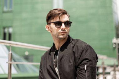 Yeşil modern binanın yakınındaki moda siyah ceket açık havada trendy güneş gözlüğü şık saç modeli ile serin genç hipster adam. Yakışıklı adam güneşli bir yaz gününde şehirde rahatlatıcı hoşlanır.