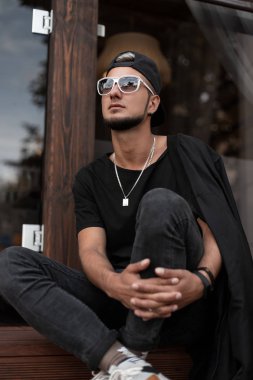 Beyaz moda güneş gözlüğü ile siyah şık t-shirt bir şık siyah kap bir sakal ile serin genç adam oturur ve bir kafede yakın merdivenlerde rüyalar. Şehir içinde açık havada kentsel hipster adam