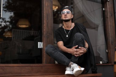 Beyaz güneş gözlüğü şık siyah giysiler içinde güneş gözlüğü trendy kap Amerikan Genç hipster adam bir Vintage binanın yakınında oturur. Yakışıklı Modern adam modeli ahşap bir merdivende dinleniyor. Erkek giyim