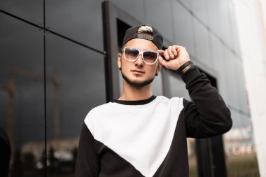 Siyah beyaz Sweatshirt şık bir siyah kap içinde bir sakal ile modern genç hipster adam standları ve açık havada gri bir duvar yakın güneş gözlüğü düzeltir. Şehirde oturan kentsel adam modeli. Sokak stili.