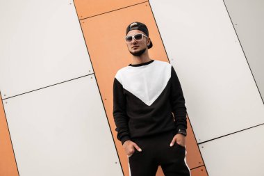 Pantolon moda Sweatshirt trendy güneş gözlüğü şık siyah kap serin genç hipster adam modern bir duvarın yakınında pozlar. Yaz gününde bir sokakta bir şehirde kentsel Amerikalı adam. Sokak stili.