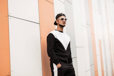 Pantolon bir moda Sweatshirt Vintage güneş gözlüğü siyah beyzbol şapkası bir sakal ile genç acımasız adam beyaz bir binanın yakınında duruyor. Bir yaz gününde şehirde modern hipster adam.