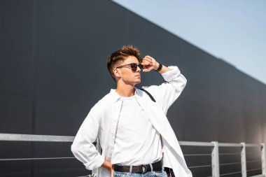 Şık genç hipster adam trendy güneş gözlüğü düzeltir. Güneşli bir günde gri bina yakın açık havada poz saç ile şık beyaz gömlek Avrupa adam modeli. Yaz yeni koleksiyon erkek giyim.