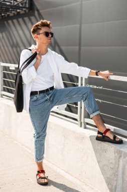 Siyah çanta ile güneş gözlüğü kırmızı Vintage sandalet trendy yaz giysileri Genç Modern adam modeli güneşli bir günde gri bir bina yakın rahatlatır. Yakışıklı adam hipster açık havada yatıyor. Şık erkek giyim.