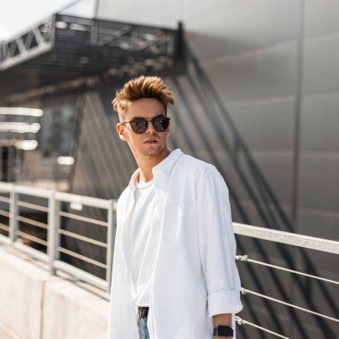 Trendy güneş gözlüğü şık bir saç modeli ile moda beyaz gömlek Avrupa Genç adam hipster güneşli bir günde sokakta gri bir binanın yakınında duruyor. Çekici adam modeli. Yazlık giysiler