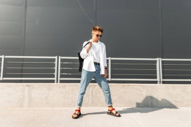 Bir bez çanta ile siyah güneş gözlüğü kırmızı Sandalet mavi kot bir Vintage gömlek kentsel hipster genç adam gri bir binanın yakınında sokakta poz veriyor. Amerikalı adam. Yaz trendy erkek giyim.