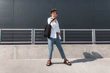 Siyah bir çanta ile koyu güneş gözlüğü mavi şık kot beyaz bir gömlek genç Avrupa adam hipster güneşli bir günde şehirde duruyor. Modern kentsel adam modeli. Şık yaz görünüm. Gençlik modası.