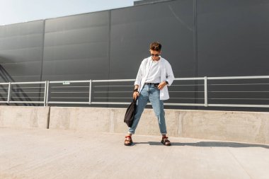 Siyah bir çanta ile güneş gözlüğü mavi şık kot beyaz bir gömlek genç Amerikan adam modeli güneşli bir günde şehir etrafında yürür. Çekici hipster adam. Şık yaz görünüm. Gençlik modası.